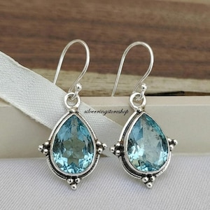 Pode incluir: Um par de brincos pendentes de prata com pedras preciosas azul-claras facetadas em forma de pera, engastadas em armações de metal polido com detalhes de contas decorativas na base e nas laterais. Os brincos pendem de ganchos de prata e incluem o texto silverringstoreshop.