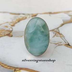 May include: Oval-shaped ring featuring a large, pale blue-green gemstone set in a silver band. The gemstone has a cloudy, marbled appearance. The text "silverringstoreshop" is visible at the bottom of the image.