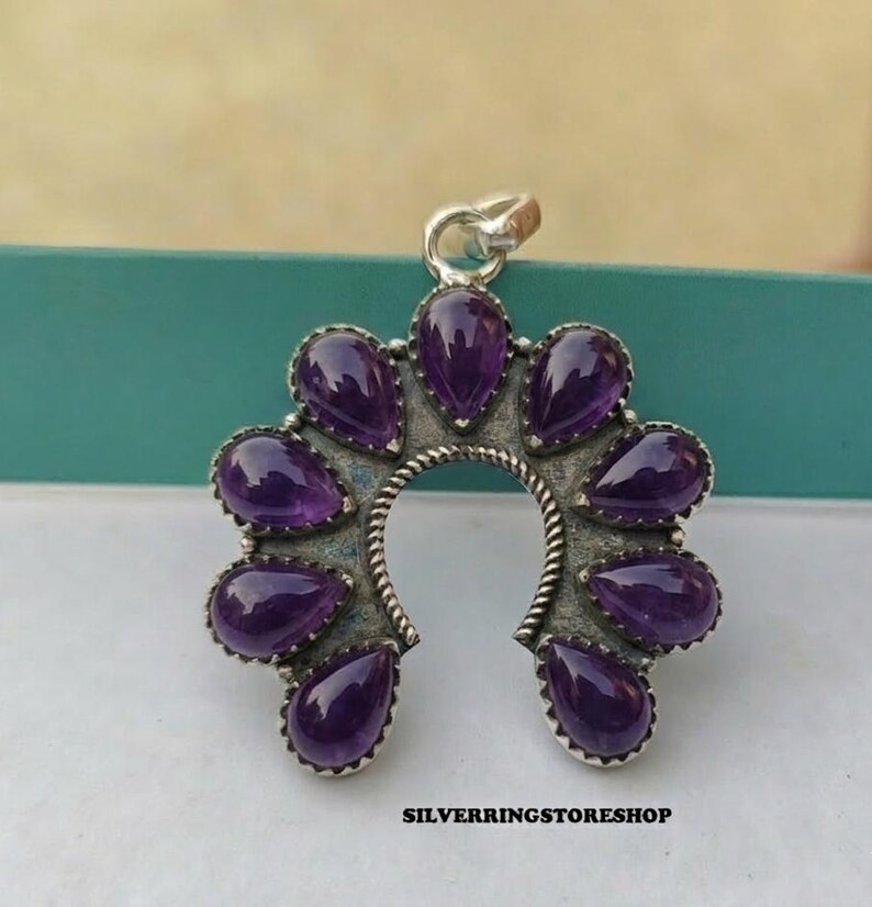 Pu&ograve; includere: Un ciondolo argentato a forma di ferro di cavallo, ornato da pietre preziose viola a forma di goccia. Il ciondolo ha un bordo strutturato e un piccolo anello per una catena. Il testo "SILVERRINGSTORESHOP" &egrave; in basso.