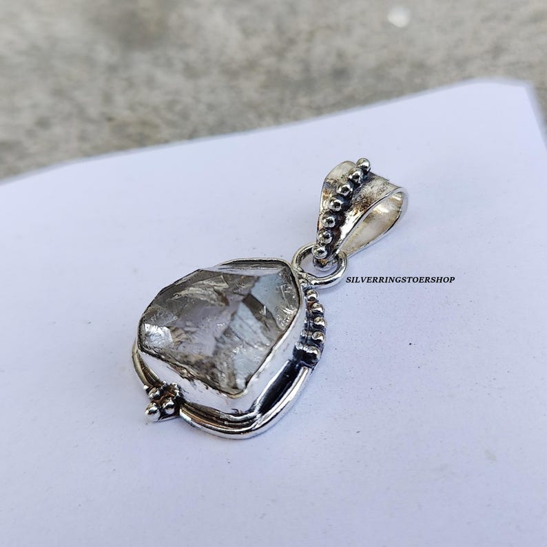 May include: A silver pendant featuring a clear, faceted quartz crystal set in a silver frame. The pendant has a beaded bail for attaching to a chain.