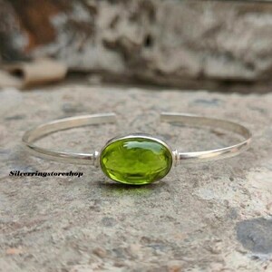 K&ouml;nnte beinhalten: Ein silbernes Armband mit einem zentralen, ovalen, durchscheinenden lindgr&uuml;nen Edelstein. Das Metallband ist glatt und poliert und weist eine &Ouml;ffnung an der R&uuml;ckseite zur Anpassung auf. Der Text "Silverringstoreshop" ist unten links zu sehen.