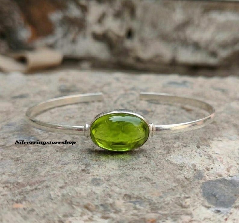 K&ouml;nnte beinhalten: Ein silbernes Armband mit einem zentralen, ovalen, durchscheinenden lindgr&uuml;nen Edelstein. Das Metallband ist glatt und poliert und weist eine &Ouml;ffnung an der R&uuml;ckseite zur Anpassung auf. Der Text "Silverringstoreshop" ist unten links zu sehen.