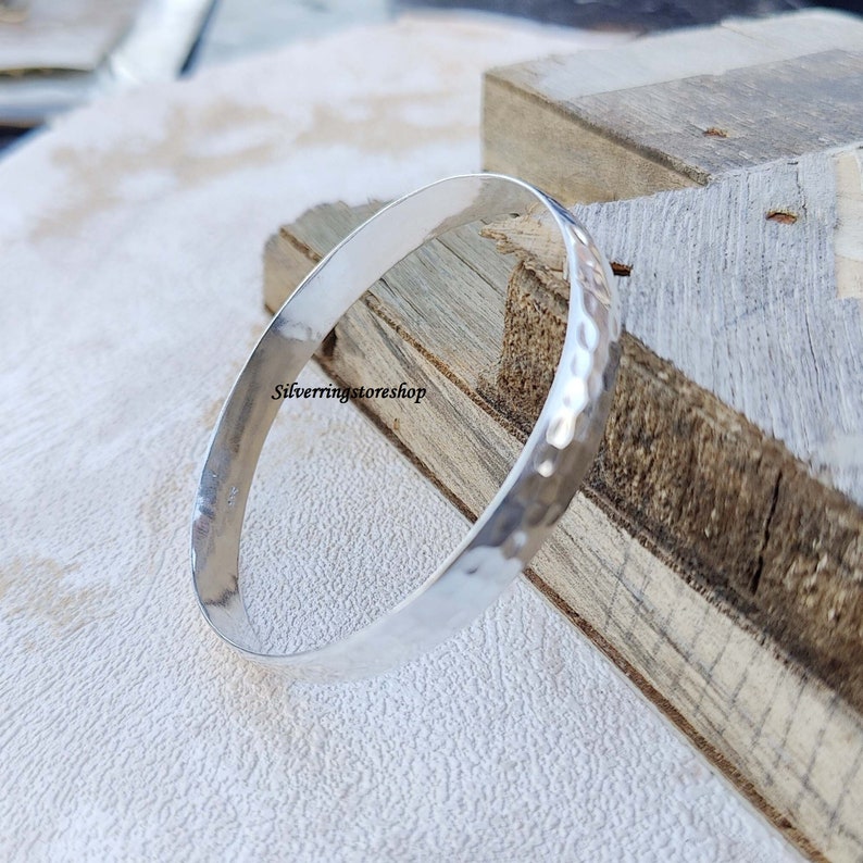 May include: A silver hammered bangle bracelet. The bracelet is smooth and shiny, with a hammered texture. The bracelet is on a wooden surface.