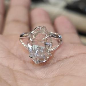 Può includere: Un anello d'argento con una punta di cristallo trasparente e sfaccettata incastonata al centro. Il cristallo è una punta naturale grezza con una forma irregolare e ruvida.