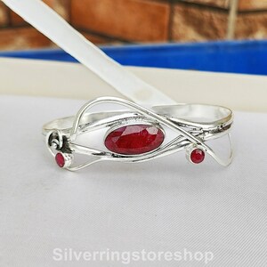 May include: A silver bracelet featuring a red ruby gemstone in the centre and two smaller red ruby gemstones on either side. The bracelet has a delicate, intricate design.