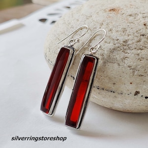 Può includere: Un paio di orecchini pendenti in argento con pietre rettangolari rosse. Gli orecchini hanno un design semplice e sono perfetti per l'uso quotidiano.