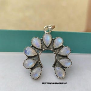 Pu&ograve; includere: Ciondolo argentato con design a ferro di cavallo, ornato da gemme di pietra di luna a forma di goccia. Le gemme hanno un'iridescenza bianca latte e blu. Il testo "SILVERRINGSTORESHOP" &egrave; in basso.
