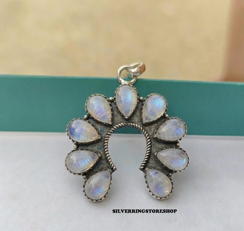 Pu&ograve; includere: Ciondolo argentato con design a ferro di cavallo, ornato da gemme di pietra di luna a forma di goccia. Le gemme hanno un'iridescenza bianca latte e blu. Il testo "SILVERRINGSTORESHOP" &egrave; in basso.