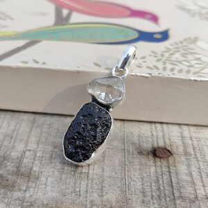 Puede incluir: Un colgante de plata con un cristal de cuarzo transparente sobre una roca volc&aacute;nica negra. El colgante est&aacute; sobre una superficie de madera.