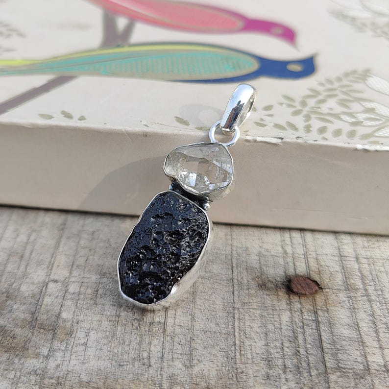 Puede incluir: Un colgante de plata con un cristal de cuarzo transparente sobre una roca volc&aacute;nica negra. El colgante est&aacute; sobre una superficie de madera.