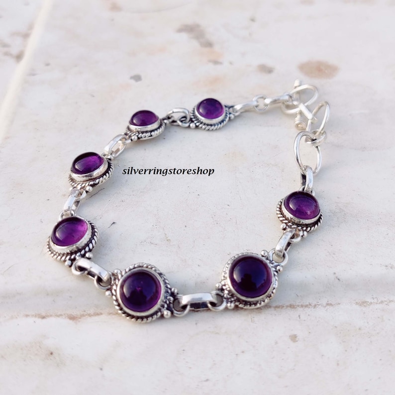 May include: A silver bracelet featuring eight round, deep purple gemstones. Each stone is set in a decorative, beaded bezel. The bracelet is connected by silver links and has a clasp closure. The text "silverringstore shop" is visible.