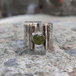 May include: A silver ring with a green gemstone set in a wide band. The band is decorated with an intricate floral pattern.