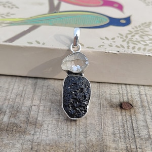 Puede incluir: Un colgante de plata con un cristal de cuarzo transparente y una piedra de obsidiana negra. El colgante est&aacute; dise&ntilde;ado en un estilo minimalista, con una simple argolla de plata.