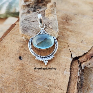 May include: A silver pendant with a light blue oval gemstone set in a decorative silver frame. The pendant has a bail for attaching to a chain.