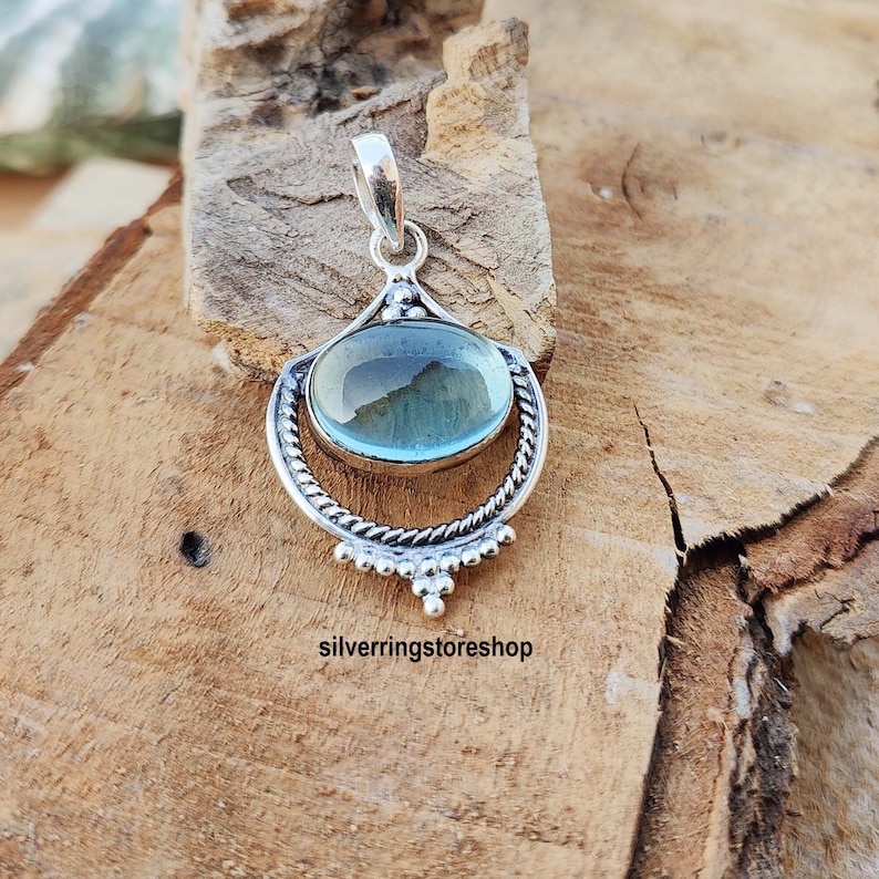 May include: A silver pendant with a light blue oval gemstone set in a decorative silver frame. The pendant has a bail for attaching to a chain.