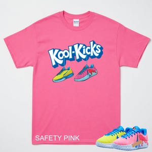 May include: A safety pink t-shirt with the text "Kool-Kicks" in blue and white. The shirt features a graphic of two trainers, one yellow and blue, the other pink and blue. The words "SAFETY PINK" are printed at the bottom.