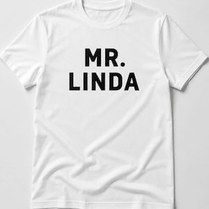 May include: A white t-shirt featuring the words "MR. LINDA" in bold, black capital letters. The shirt has a crew neck and short sleeves. The text is centrally positioned on the front of the garment.