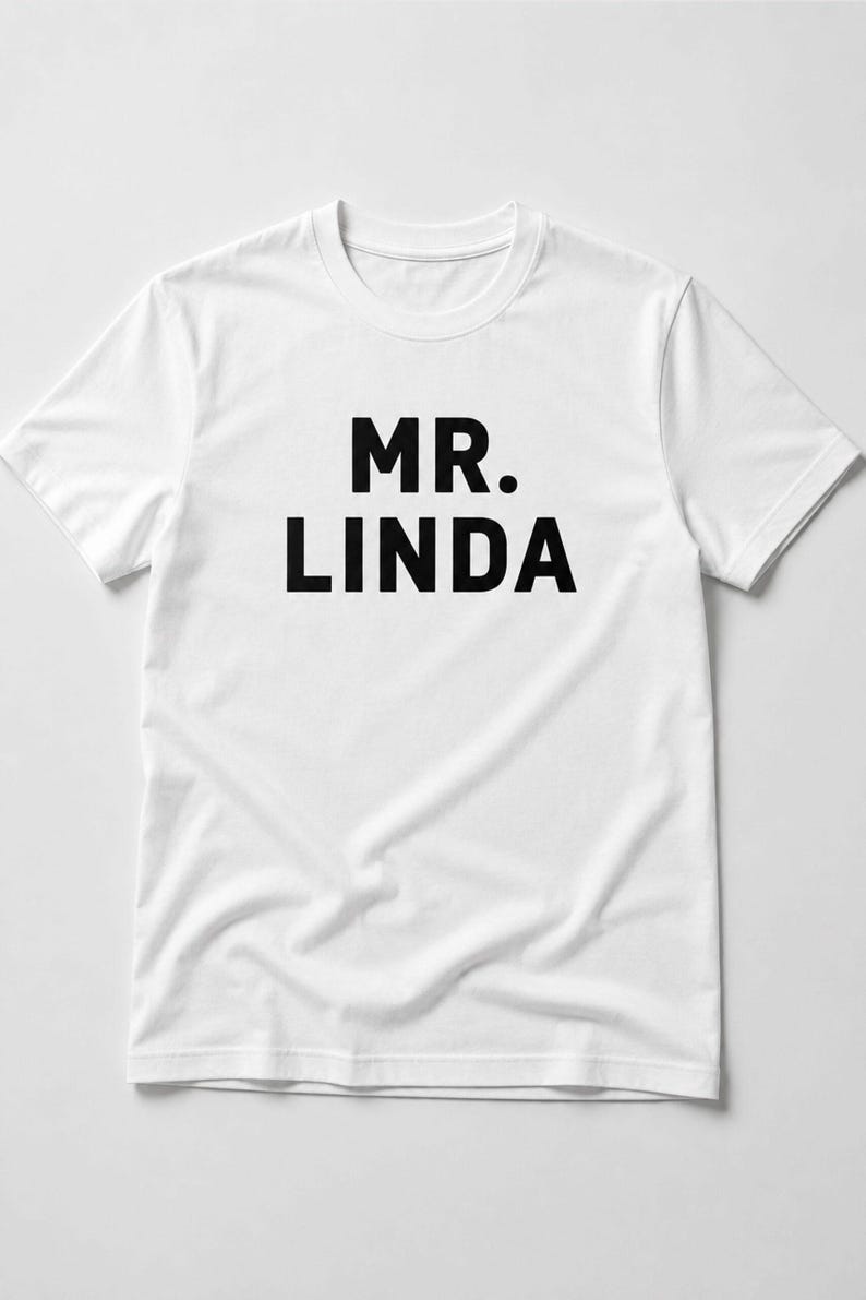 May include: A white t-shirt featuring the words "MR. LINDA" in bold, black capital letters. The shirt has a crew neck and short sleeves. The text is centrally positioned on the front of the garment.