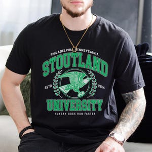 May include: Black t-shirt with a green and white graphic print of a bird and the text "Stoutland University, Philadelphia Pennsylvania, Est. 1984, Hungry Dogs Run Faster".