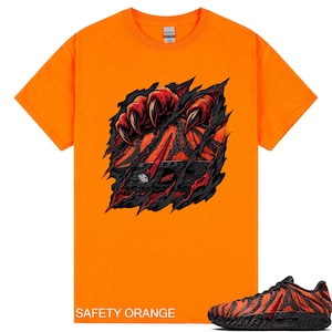 以下が含まれることがあります： 鮮やかなセーフティオレンジ色の半袖Tシャツ。大きな赤い爪が生地を引き裂き、黒と赤の模様が入ったスニーカーが現れるグラフィックが描かれています。スニーカーのグラフィックには赤色でNIKEという文字がプリントされています。