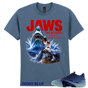 Pode incluir: Uma t-shirt de manga curta azul ardósia com um gráfico de um tubarão a emergir da água atrás de um jogador de basquetebol num cais de madeira. O texto diz JAWS THE REVENGE This Time It's Personal. Abaixo, um ténis desportivo azul escuro e claro.