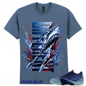 Puede incluir: Una camiseta azul pizarra con un gráfico de un tiburón emergiendo de la palabra DIFFERENT, rodeado de salpicaduras de pintura azul y roja. El texto INDIO BLUE está impreso en la esquina inferior izquierda, junto a una zapatilla deportiva azul.