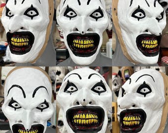 May include: A collection of six white clown masks with black accents. Each mask features exaggerated black eyebrows, eye details, and a wide, grinning mouth revealing yellow teeth. The masks have a beige backing.