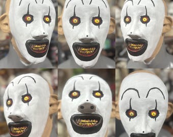 May include: A collection of six white clown masks with black and red details. Each mask features a wide, menacing grin with gold teeth, red eyes, and black eyebrows. The masks have large, exaggerated ears and are designed for Halloween or costume use.