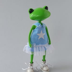 Puede incluir: Una figura de rana verde con un vestido azul claro con una estrella blanca y una falda de tul blanco. La rana tiene ojos negros y zapatillas blancas con cordones. La figura está sobre una superficie blanca.