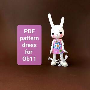 May include: PDF pattern dress for Ob11 doll. The doll is wearing a pink dress with a floral print and white shoes. The doll has white bunny ears and a white face with red cheeks.