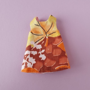 Może przedstawiać: Sukienka bez rękawów z kwiatowym nadrukiem w odcieniach pomarańczowego, brązowego i żółtego. Sukienka ma prosty krój i idealnie nadaje się dla lalki.