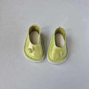 May include: A pair of small, light green doll shoes. The shoes have a rounded toe, a decorative embellishment, and a white sole. The shoes are made of a shiny material and are likely for a doll or other small figure.