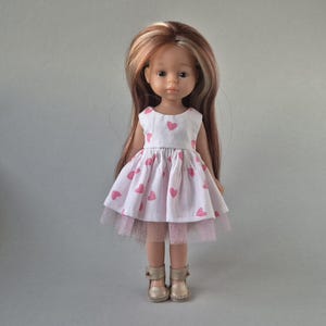 Peut inclure: Une poupée aux longs cheveux bruns avec des mèches blondes, vêtue d'une robe sans manches blanche à motif de cœurs roses avec un ourlet en tulle rose clair. La poupée porte également des sandales dorées métallisées avec une boucle.