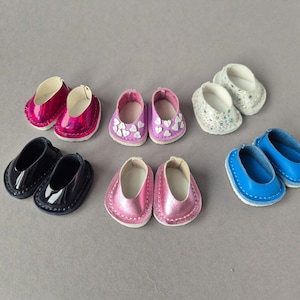 May include: A collection of miniature doll shoes in various colors and styles. The shoes include pairs in shiny pink, black patent leather, lavender with heart accents, white with glitter, pink, and blue. The shoes are arranged on a gray surface.