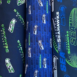 May include: Three different fabric patterns featuring the Seattle Seahawks logo. The first pattern is a dark blue background with a camouflage green and white Seahawks logo. The second pattern is a dark blue background with a white Seahawks logo. The third pattern is a dark blue background with a green and white Seahawks logo and a graphic of the Seattle skyline.