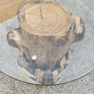 Pode incluir: Uma mesa de madeira rústica com acabamento de grão de madeira natural. A mesa é feita de um tronco de árvore e tem uma superfície áspera e irregular. A mesa é apoiada por uma base de vidro.
