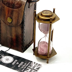 May include: A brass hourglass with pink sand inside, in a brown leather carrying case. The case has a strap and a brass buckle. The hourglass is 3 inches tall and the case is 6 inches tall.