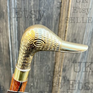 Può includere: Un bastone da passeggio in legno con un manico a testa di anatra in ottone. Il bastone ha un fusto in legno marrone e una fascia in ottone attorno alla parte superiore. Un'etichetta attaccata al bastone recita "UK Etsy Seller Delivery with in 3 days www.ministryofantiques.etsy.com".