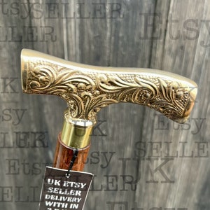 May include: A wooden walking stick with a gold-toned metal handle featuring an ornate floral design. The handle is T-shaped and has a brass cap on the end. The stick is labeled "UK Etsy Seller Delivery within 3 days" and "www.ministryofantiques.etsy.com".