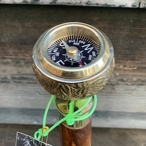 Peut inclure: Une canne de marche en bois avec un compas en laiton sur le dessus. Le cadran du compas est noir avec des marques blanches et une aiguille rouge. Le bâton a un motif de grain de bois brun et noir. Le bâton a une sangle verte attachée à lui avec une épingle de sûreté.