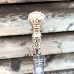 May include: A silver walking stick with an ornate, engraved handle. The stick is black and has a silver band around it. The text on the tag reads "DELIVERY WITH IN 3 DAYS".