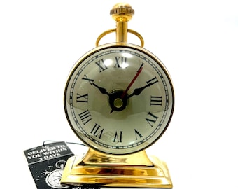 May include: A gold-toned tabletop clock with Roman numeral markings on the face. The clock has a round face and a decorative base.