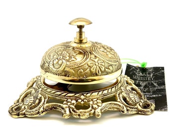 May include: A gold-toned metal bell with an ornate design. The bell is on a base with a decorative design. The bell is labeled "Ministry of Antiques Teller Dispatch".