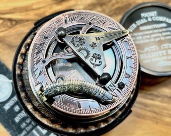May include: A vintage-style sundial and compass set. The sundial features a copper-toned face with intricate markings and a gnomon. The compass is in a round, dark-colored case. The set is presented on a wooden surface, with the text "ELLIOTT BRO" visible.