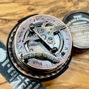 May include: A vintage-style sundial and compass set. The sundial features a copper-toned face with intricate markings and a gnomon. The compass is in a round, dark-colored case. The set is presented on a wooden surface, with the text "ELLIOTT BRO" visible.