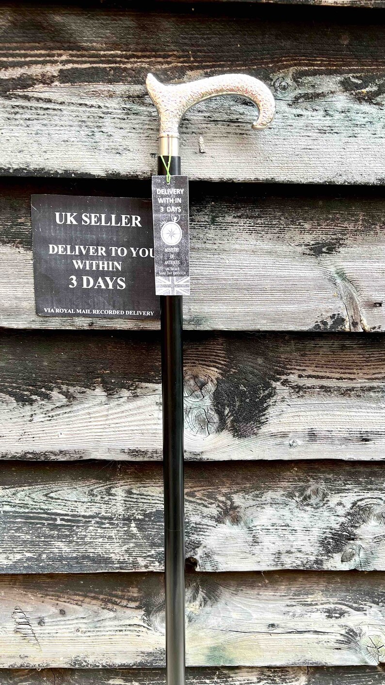 K&ouml;nnte beinhalten: Ein schwarzer Gehstock mit einem silbernen Griff. Der Griff hat ein dekoratives Design. Der Stock lehnt an einer Holzwand. Der Text auf dem Schild lautet "UK Seller Deliver to you within 3 days via Royal Mail Recorded Delivery".