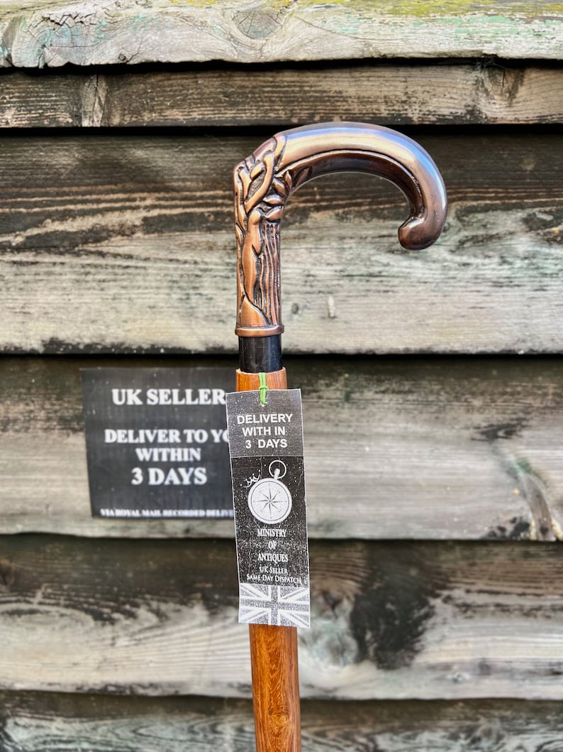 May include: A wooden walking stick with a copper-colored handle featuring a tree design. The handle has a curved top and a black metal band. A tag attached to the stick reads "Delivery with in 3 days" and "Ministry of Antiques UK Seller Same Day Dispatch".