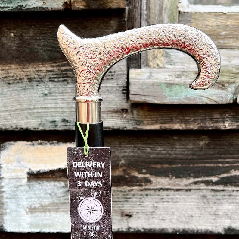 K&ouml;nnte beinhalten: Ein silberner Gehstock mit einem verzierten Design. Der Stock hat einen schwarzen Griff und eine silberne Spitze. Die Spitze des Stocks ist hakenf&ouml;rmig. Der Stock h&auml;ngt an einer Holzwand. Ein Schild ist am Stock befestigt, auf dem "Lieferung innerhalb von 3 Tagen" steht.