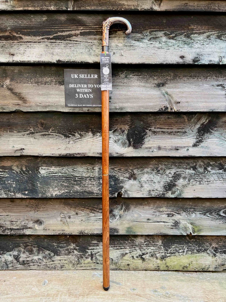 May include: A wooden walking stick with a silver handle featuring a floral design. The stick has a brown finish and a black rubber tip. The stick is leaning against a wooden wall.