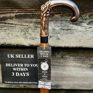 May include: A wooden walking stick with a copper-colored handle featuring a detailed design. The handle is curved and has a decorative design. The stick is brown and has a natural wood grain. The stick is accompanied by a tag that reads "Delivery within 3 days" and "Ministry of Antiques UK Seller Same Day Dispatch".
